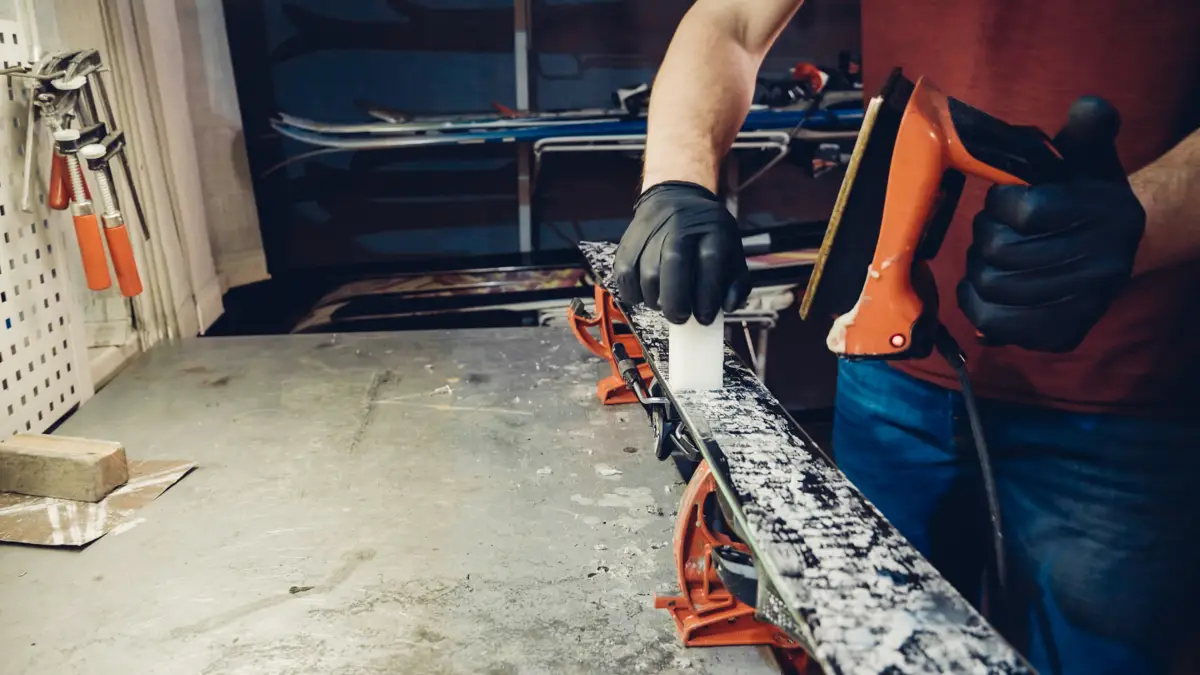 Un individu ganté applique de la cire sur un ski noir et blanc avec un fer orange sur un établi métallique.