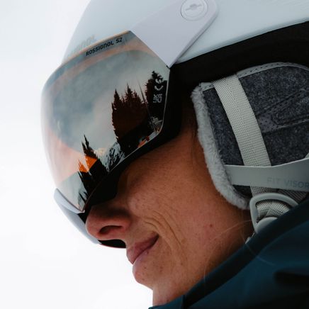 Reflets d'arbres et de ciel orangé dans la visière miroir du casque blanc d'une personne souriante.