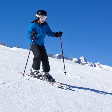 Skieur en veste bleue et casque noir dévale une piste enneigée sous un ciel bleu éclatant, avec des montagnes en fond.