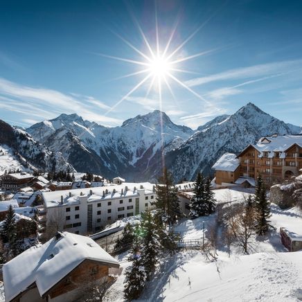 Paysage hivernal montagnard. Un village enneigé sous un soleil radieux, entouré de sommets alpins majestueux et d'un ciel bleu.